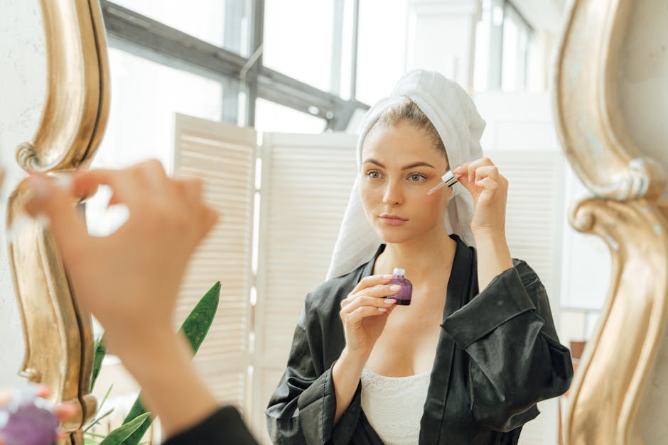 A woman in a towel and robe applies skincare serum with a dropper in front of a mirror