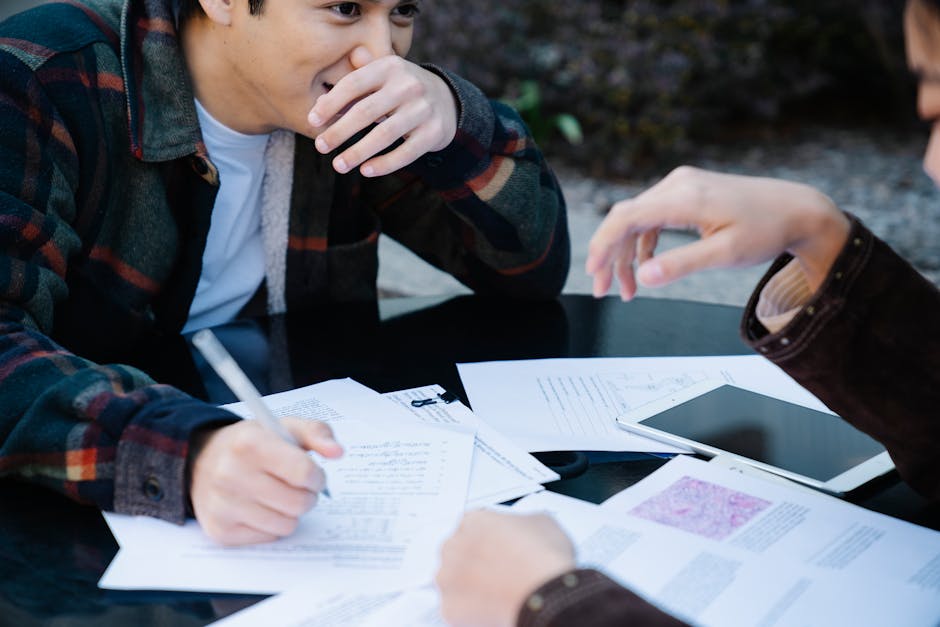 Two students sharing ideas over study papers and a tablet outdoors.
