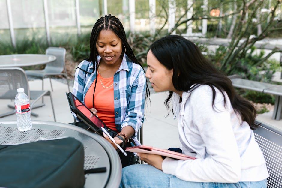 Two young women studying together using a tablet at outdoor campus seating.